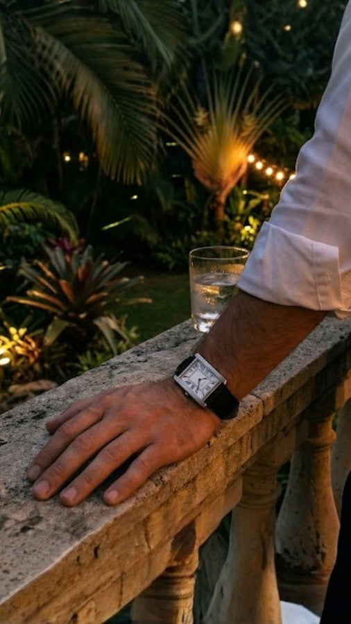 Relógio retangular prateado de pulseira preta no pulso, com a mão apoiada em balaustrada e vegetação ao fundo.