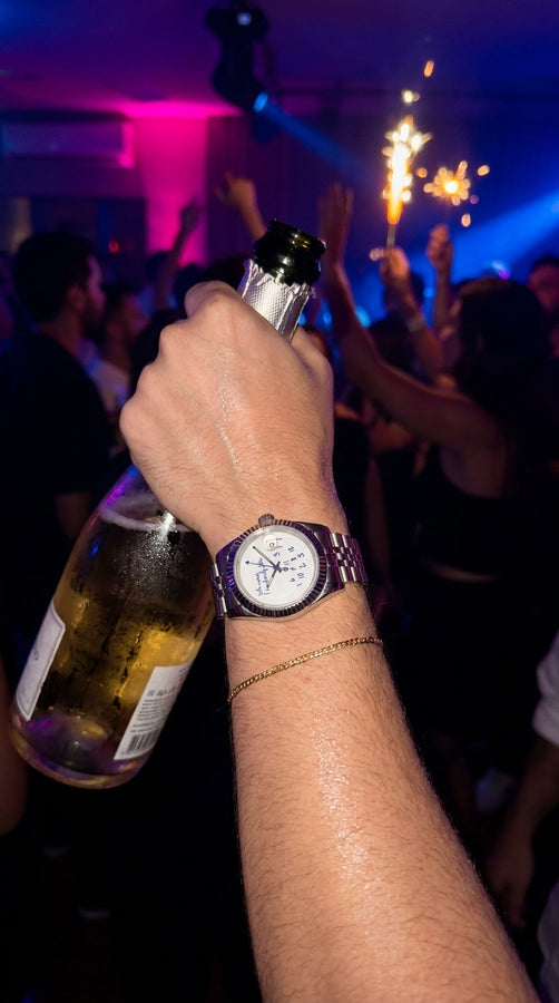 Pessoa com relógio de mostrador branco e pulseira preta segurando uma garrafa em uma festa.