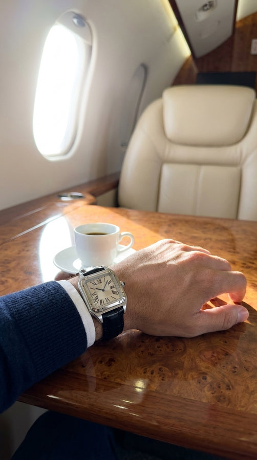 Relógio retangular prateado de pulseira preta no pulso, com o braço apoiado em uma mesa junto à janela de avião.