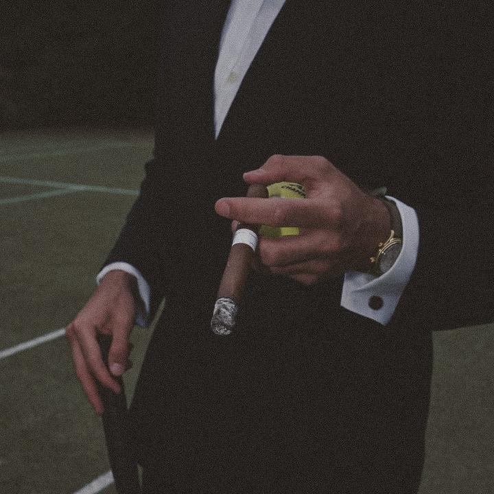Um homem vestido com um smoking elegante segura um charuto e uma bola de tênis, exibindo um relógio de luxo no pulso. Ele está em um ambiente sofisticado, com o foco no estilo de vida luxuoso e no uso de acessórios finos.