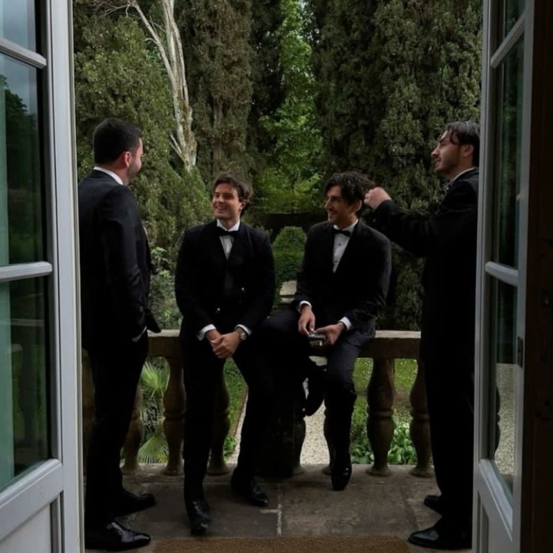 Quatro homens com smoking sentados e conversando em um ambiente sofisticado, com vista para um jardim bonito. Eles estão em um cenário elegante, com relógios luxuosos visíveis em seus pulsos.