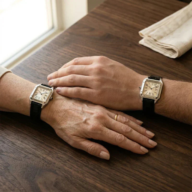 Duas mãos sobre uma mesa de madeira usando relógios retangulares com pulseiras pretas, em imagem para o Dia das Mães.