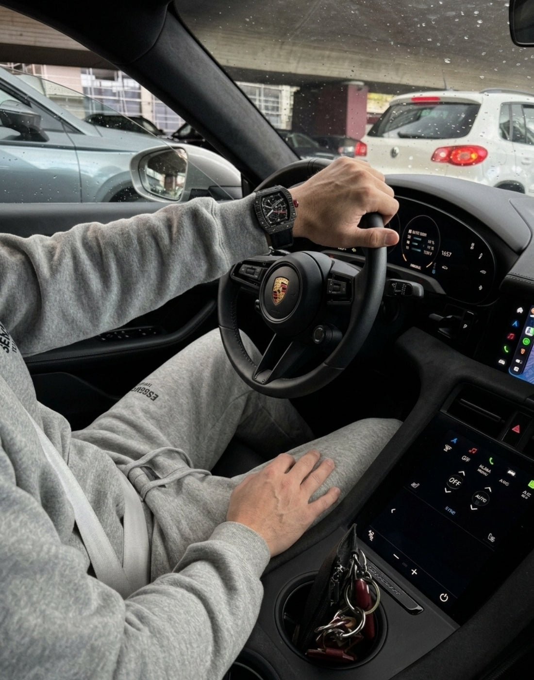 Relógio esportivo escuro no pulso de um motorista com roupa cinza, segurando o volante de um carro.