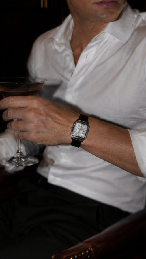 Homem de camisa branca usando relógio retangular de pulseira preta e segurando um copo.