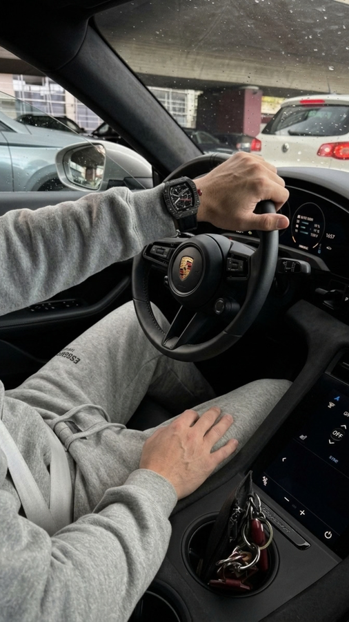 Vista do interior de um carro, com relógio escuro no pulso do motorista enquanto segura o volante.