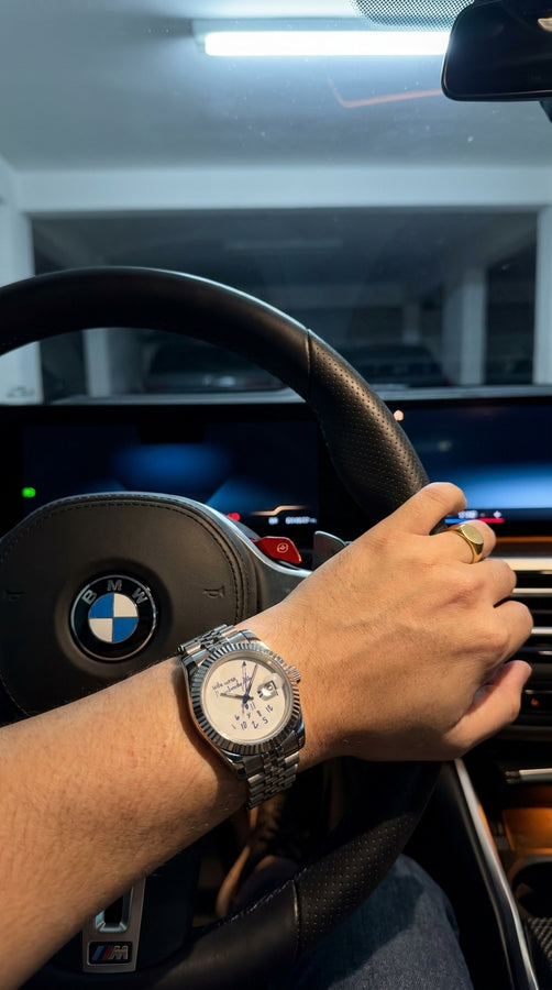 Relógio prateado de mostrador claro no pulso de uma pessoa segurando o volante de um carro.