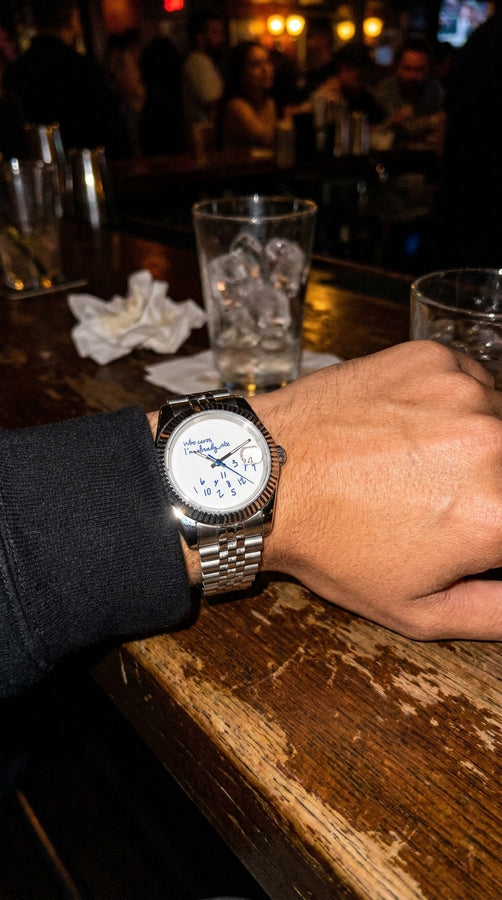 Relógio prateado de mostrador branco e pulseira metálica no pulso, apoiado em mesa de restaurante.